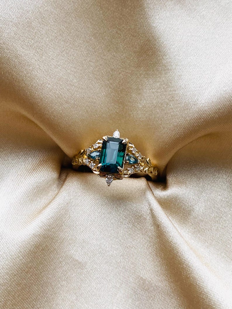 May include: A gold ring with a large emerald green gemstone surrounded by smaller diamonds. The ring is set on a white satin background.