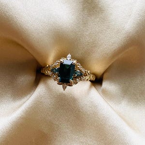 May include: A gold ring with a large emerald-cut green gemstone surrounded by smaller diamonds. The ring has a delicate floral design.
