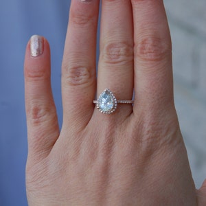 May include: A pear-shaped diamond engagement ring with a halo setting in a rose gold band. The ring is on a person's finger.
