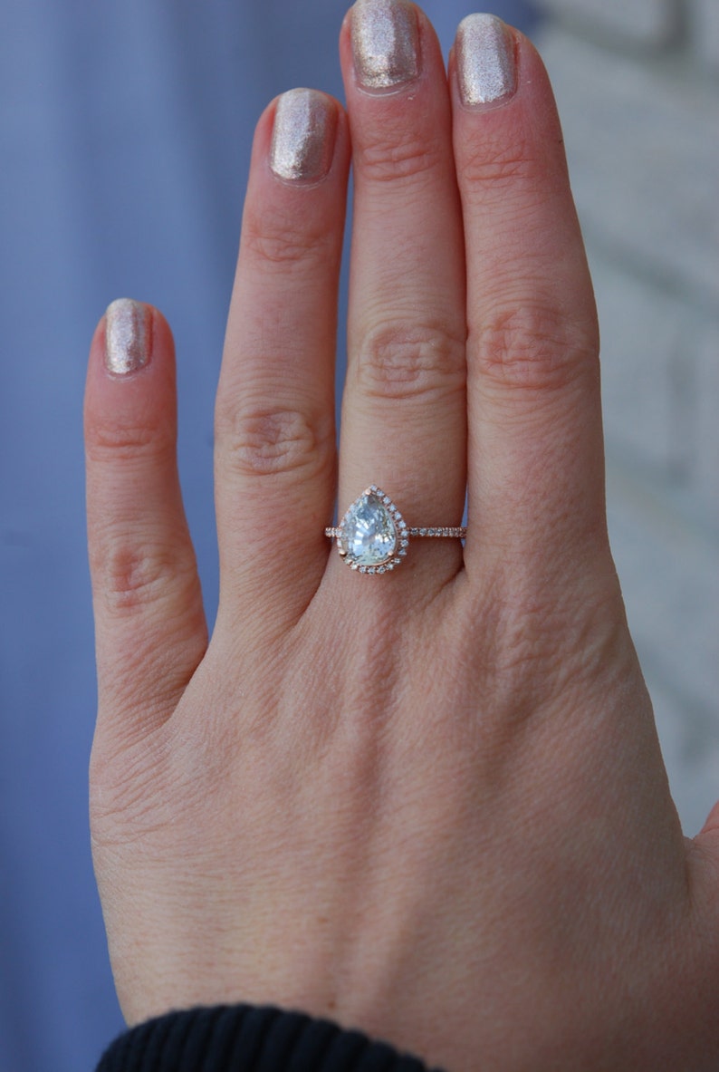 May include: A pear-shaped diamond engagement ring with a halo setting in a rose gold band. The ring is on a person's finger.