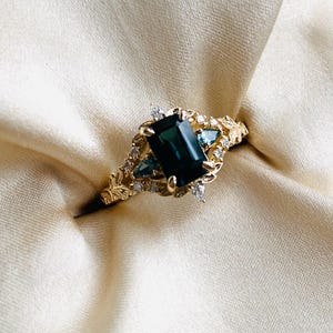 May include: A gold ring with a dark green emerald-cut gemstone surrounded by smaller diamonds and a leafy design.