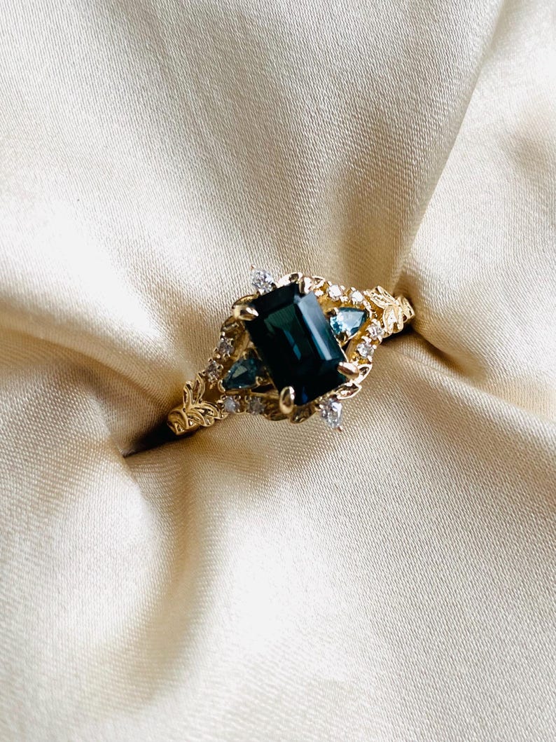 May include: A gold ring with a dark green emerald-cut gemstone surrounded by smaller diamonds and a leafy design.