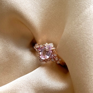 Puede incluir: Un delicado anillo de oro rosa con una gran piedra ovalada rosa rodeada de piedras más pequeñas de color rosa y blanco. El anillo está colocado sobre un fondo de satén blanco.