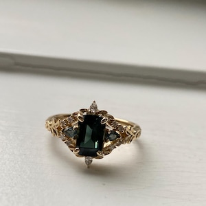 May include: A gold ring with a dark green gemstone surrounded by smaller clear gemstones. The ring has a delicate floral design.