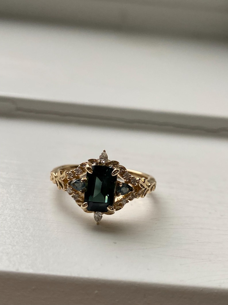 May include: A gold ring with a dark green gemstone surrounded by smaller clear gemstones. The ring has a delicate floral design.