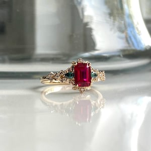 May include: A gold ring featuring a large emerald-cut ruby, surrounded by smaller diamonds and green stones. The ring has a delicate floral design.