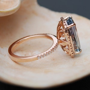 May include: A rose gold engagement ring with a large, rectangular, blue gemstone set in a delicate, ornate halo setting. The band is adorned with a row of small, sparkling diamonds.