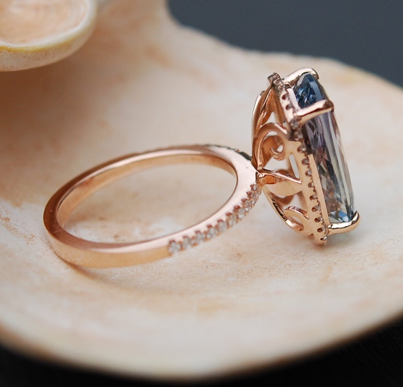 May include: A rose gold engagement ring with a large, rectangular, blue gemstone set in a delicate, ornate halo setting. The band is adorned with a row of small, sparkling diamonds.