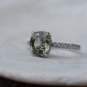 May include: A close-up of a ring featuring a large, oval-cut, olive-green gemstone. The ring has a silver band with small, clear stones set along the top half. The gemstone is held in place by four prongs.