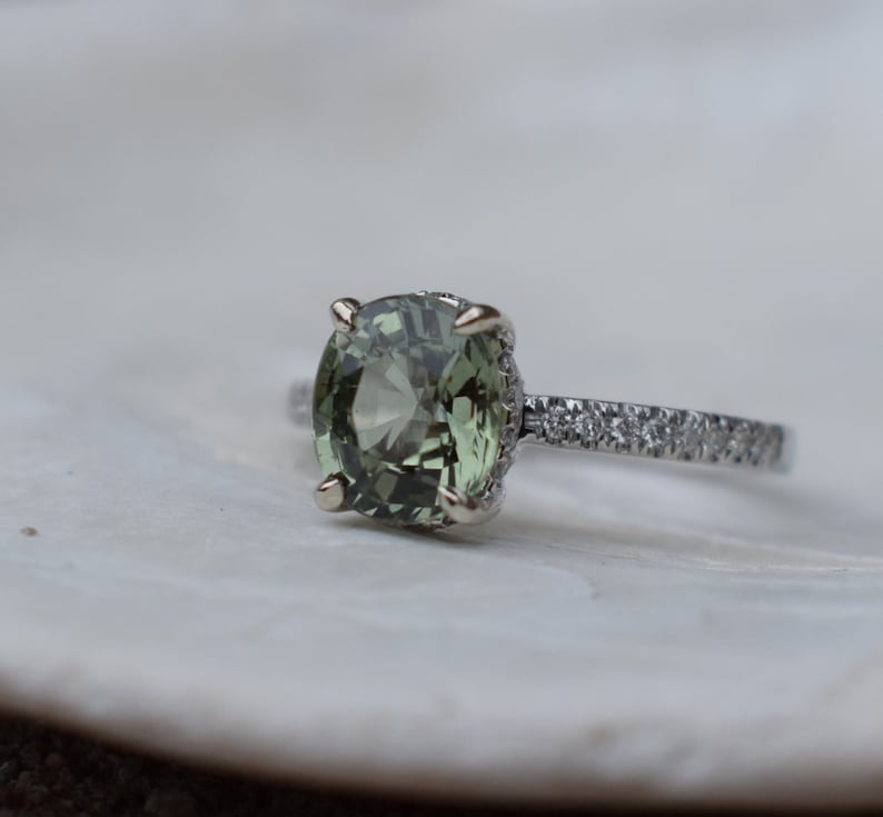 May include: A close-up of a ring featuring a large, oval-cut, olive-green gemstone. The ring has a silver band with small, clear stones set along the top half. The gemstone is held in place by four prongs.