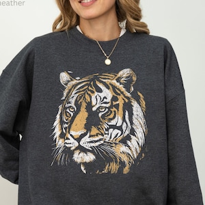 Op de afbeelding: Donkergrijze sweatshirt met een gedetailleerde tijgergezichtsafbeelding in tinten oranje, wit en zwart. De sweatshirt heeft een ronde hals en lange mouwen. De tekst "MADE WITH LOVE BY ANIMAL PARC APPAREL" staat rechtsonder.