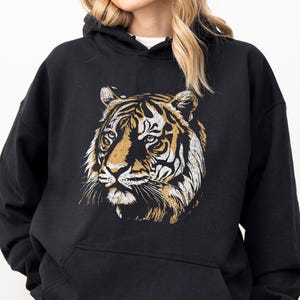 May include: Black hooded sweatshirt featuring a detailed tiger face graphic in shades of gold, white, and brown. The sweatshirt has a kangaroo pocket and a drawstring hood. The text "COLOR: BLACK" is at the bottom left.