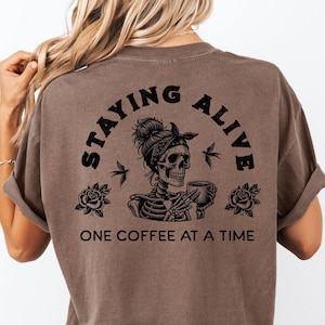 Op de afbeelding: Espresso-kleurig t-shirt met een afbeelding van een skelet met een bandana en een koffiekopje, met de tekst "STAYING ALIVE" en "ONE COFFEE AT A TIME". Het shirt is van Animal Parc Apparel.