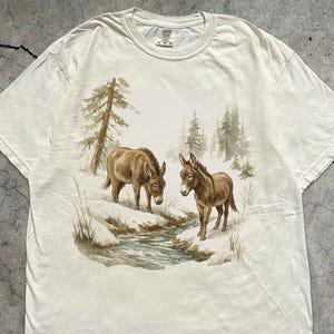 Puede incluir: Camiseta de color marfil con una ilustración en acuarela de dos burros en un paisaje nevado con un arroyo y árboles. La camiseta tiene cuello redondo y mangas cortas. El texto "MADE WITH LOVE BY ANIMAL PARC APPAREL" está impreso en la parte inferior derecha.
