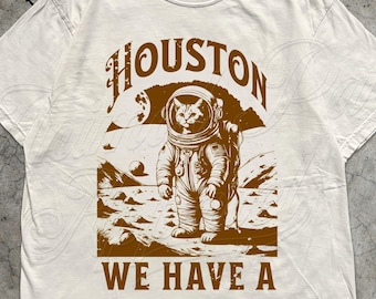 May include: Ivory-colored t-shirt featuring a brown graphic of a cat in an astronaut suit on a planet surface. The text "HOUSTON WE HAVE A KITTEN" is above and below the image. The shirt is made with love by Animal Parc Apparel.