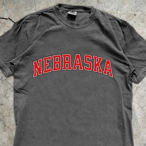 Op de afbeelding: Een grijs t-shirt met het woord "NEBRASKA" in rood, gebogen over de borst. Het shirt heeft een vintage, versleten uitstraling. Een banner rechtsonder zegt "MADE WITH LOVE BY ANIMAL PARC APPAREL".