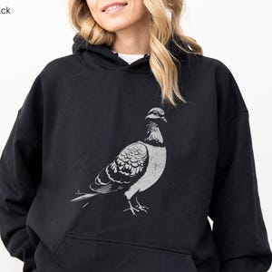 May include: Black hoodie featuring a detailed gray illustration of a pigeon. The bird is depicted in a side profile, showcasing its feathers and form. The hoodie has a kangaroo pocket and a drawstring hood. The text "MADE WITH LOVE BY ANIMAL PARC APPAREL" is printed on a banner.