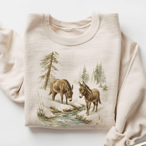 May include: Sand-coloured sweatshirt with a watercolour illustration of two donkeys in a snowy landscape with a stream and pine trees. The text "MADE WITH LOVE BY ANIMAL PARC APPAREL" is printed on the bottom right.