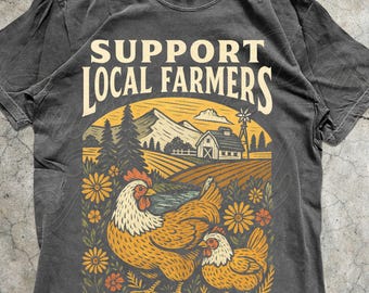 May include: Pepper-coloured t-shirt with a graphic supporting local farmers. The design features a farm scene with chickens, flowers, and the text "SUPPORT LOCAL FARMERS." The shirt is from Animal Parc Apparel.