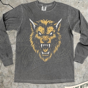 May include: A charcoal gray long-sleeve shirt featuring a graphic of a snarling wolf head in yellow and white. The wolf has bared teeth and pointed ears. The shirt has a crew neckline and a tag at the collar that reads Comfort Colors.