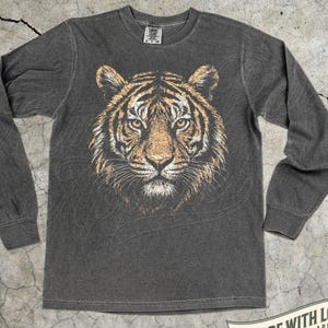 May include: Pepper-colored long-sleeve shirt featuring a detailed tiger face graphic. The tiger's face is rendered in shades of brown, orange, and white, with a focused gaze. The shirt includes the text "MADE WITH LOVE BY ANIMAL PARC APPAREL".