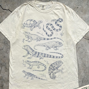 May include: Ivory t-shirt with a blue illustration of reptiles such as a turtle, snake, iguanas, and chameleons. The shirt has a crew neck and short sleeves. The text "MADE WITH LOVE BY ANIMAL PARC APPAREL" is printed on a banner. The color is ivory.