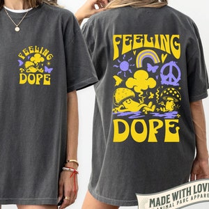 May include: A dark grey t-shirt with a yellow and purple graphic print that says "Feeling Dope" on the front and back. The back graphic features a mushroom, a rainbow, a peace sign, and a sun.