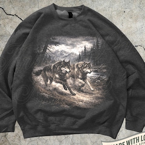 Pode incluir: Camisola cinza escuro com uma ilustração detalhada de dois lobos correndo por uma paisagem florestal. O design inclui árvores, montanhas e um rio. O texto "MADE WITH LOVE BY ANIMAL PARC APPAREL" também é visível.