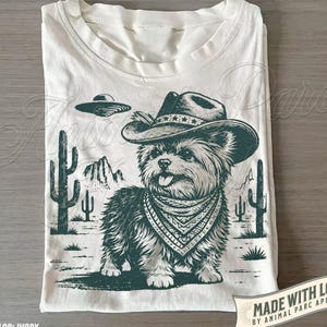 Pode incluir: Uma t-shirt cor de marfim com um gráfico verde-escuro de um cão felpudo a usar um chapéu de cowboy e uma bandana estampada. A cena inclui uma paisagem desértica com catos, montanhas e um disco voador sobre o cão.