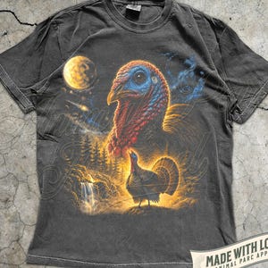 Puede incluir: Una camiseta de manga corta gris carb&oacute;n con un estampado gr&aacute;fico de una cabeza de pavo grande y un pavo m&aacute;s peque&ntilde;o en un paisaje forestal con una cascada, una luna amarilla brillante y una estrella fugaz sobre un fondo c&oacute;smico oscuro.