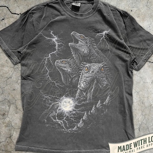 Puede incluir: Camiseta color pimienta con una ilustración detallada de tres iguanas con la boca abierta, sobre un fondo de relámpagos y luna llena. La camiseta incluye el texto "MADE WITH LOVE BY ANIMAL PARC APPAREL".