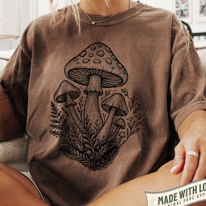 Könnte beinhalten: Espressofarbenes T-Shirt mit einer detaillierten Illustration von Pilzen und botanischen Elementen. Die Grafik ist in Schwarz und Weiß gehalten. Das Shirt hat eine lockere Passform und kurze Ärmel. Der Text "MADE WITH LOVE" ist unten rechts sichtbar.