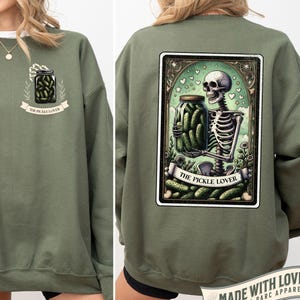 May include: Olive green sweatshirt with a pickle-themed design. The front features a jar of pickles with the text 'THE PICKLE LOVER'. The back displays a tarot card design with a skeleton holding a pickle jar, surrounded by pickles and the same text. The design is framed with a decorative border.