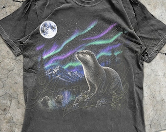 May include: Pepper-colored t-shirt featuring a detailed graphic of an otter gazing at the aurora borealis and a full moon. The design includes mountains and stars, with the text "MADE WITH LOVE BY ANIMAL PARC APPAREL" on a banner.