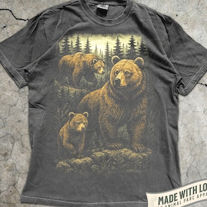 May include: Pepper-colored t-shirt featuring a detailed illustration of three brown bears in a forest setting. The design includes a mother bear and two cubs, with a backdrop of pine trees. The shirt has a classic crew neck and short sleeves.
