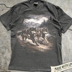K&ouml;nnte beinhalten: Pfefferfarbenes T-Shirt mit einer detaillierten Grafik von zwei W&ouml;lfen, die durch eine Waldlandschaft mit Bergen im Hintergrund laufen. Das Shirt hat einen klassischen Rundhalsausschnitt und kurze &Auml;rmel. Der Text "MADE WITH LOVE BY ANIMAL PARC APPAREL" ist unten rechts aufgedruckt.