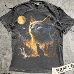 May include: A charcoal grey short-sleeve t-shirt featuring a graphic print of a large cat face looking towards a full moon and a shooting star above a smaller cat sitting in a forest landscape with a waterfall. The shirt has a crew neckline.