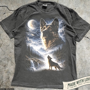 以下が含まれることがあります： ペッパーカラーのTシャツ。ドイツシェパード犬と遠吠えするオオカミの詳細なグラフィックが、満月の下の山と滝の背景に描かれています。シャツには「MADE WITH LOVE BY ANIMAL PARC APPAREL」の文字が入っています。