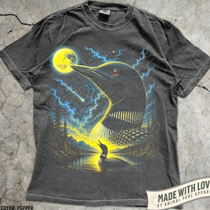 May include: A charcoal grey t-shirt featuring a graphic print of a loon bird against a night sky with a yellow moon, blue lightning, and a smaller loon swimming on a lake. The shirt has a crew neckline and short sleeves with a vintage wash finish.