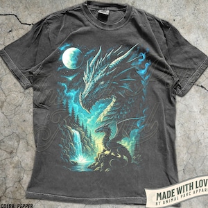 May include: Pepper-colored t-shirt featuring a detailed dragon graphic. The design includes a large dragon's head, a smaller dragon, a waterfall, and a full moon. The text "MADE WITH LOVE BY ANIMAL PARC APPAREL" is printed on a banner.