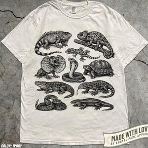 Puede incluir: Una camiseta de manga corta color marfil con un estampado gráfico en tinta negra de varios reptiles y anfibios, incluyendo una iguana, un camaleón, un lagarto de cuello, una cobra, una tortuga, un caimán y serpientes en la parte frontal.