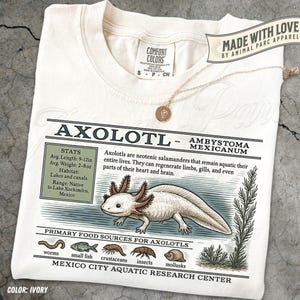 May include: Cream-coloured t-shirt with an axolotl graphic and scientific information. The design includes the axolotl's stats, habitat, and food sources. The shirt also has the text "MADE WITH LOVE BY ANIMAL PARC APPAREL". A gold necklace with a pendant is draped over the shirt.