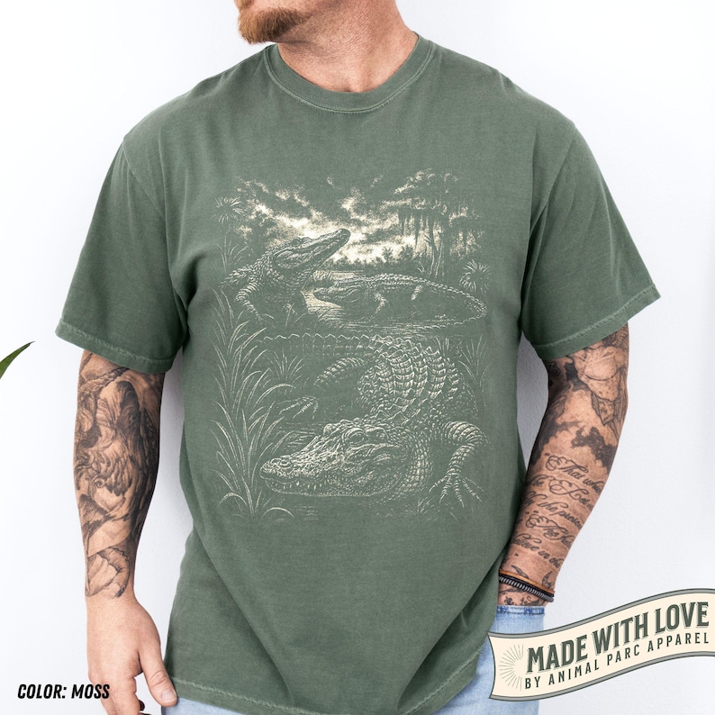 May include: Moss green t-shirt featuring a detailed illustration of alligators in a swamp setting. The design includes multiple alligators, water, and vegetation. The text "MADE WITH LOVE BY ANIMAL PARC APPAREL" is printed on a banner at the bottom right.