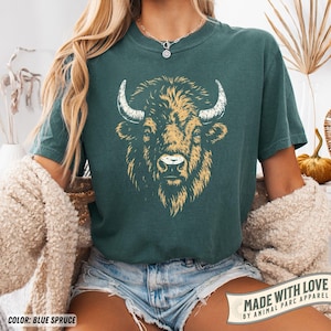 May include: Blue spruce t-shirt featuring a detailed illustration of a bison's head with tan and white accents. The shirt has short sleeves and a crew neck. The text "MADE WITH LOVE BY ANIMAL PARC APPAREL" is printed on a banner.