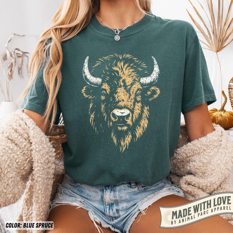 May include: Blue spruce t-shirt featuring a detailed illustration of a bison's head with tan and white accents. The shirt has short sleeves and a crew neck. The text "MADE WITH LOVE BY ANIMAL PARC APPAREL" is printed on a banner.