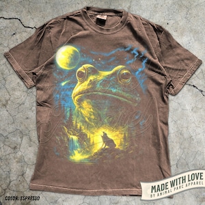 May include: A brown short-sleeve t-shirt featuring a graphic print of a large frog head above a landscape with a waterfall, a small wolf silhouette, and a bright yellow moon in a starry blue sky. The shirt has a crew neckline and a relaxed fit.