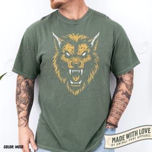 May include: Moss green t-shirt with a distressed yellow and white wolf graphic. The wolf has sharp teeth and is snarling. The shirt has short sleeves and a crew neck. The text "MADE WITH LOVE BY ANIMAL PARC APPAREL" is printed on a banner at the bottom.