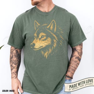 May include: Moss green t-shirt featuring a detailed illustration of a wolf's head in gold and black. The shirt has a classic crew neck and short sleeves. The text "MADE WITH LOVE BY ANIMAL PARC APPAREL" is printed on a white banner.