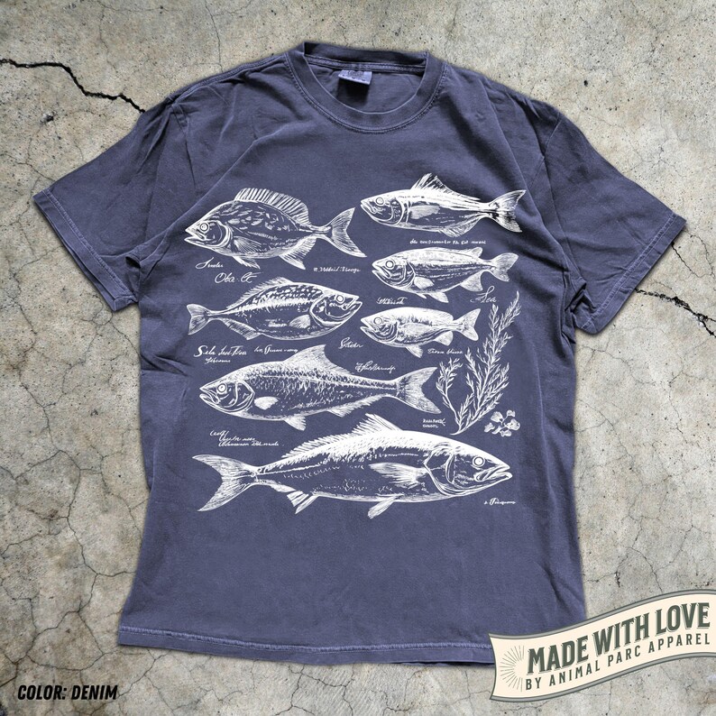 May include: Denim blue t-shirt featuring a detailed white illustration of various fish species, each labeled with their scientific names. The shirt includes the text "MADE WITH LOVE BY ANIMAL PARC APPAREL" on a banner.