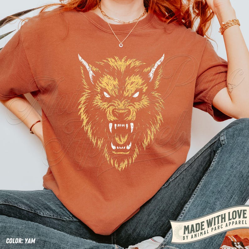 May include: A yam-coloured t-shirt with a detailed illustration of a snarling wolf's head in yellow and white. The shirt has a relaxed fit and is paired with blue jeans. The text "MADE WITH LOVE BY ANIMAL PARC APPAREL" is visible.